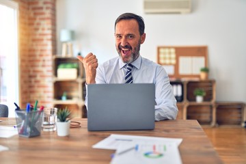 Obraz premium Middle age handsome businessman wearing tie sitting using laptop at the office smiling with happy face looking and pointing to the side with thumb up.