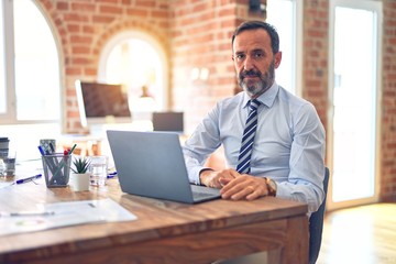 Middle age handsome businessman wearing tie sitting using laptop at the office with serious expression on face. Simple and natural looking at the camera.