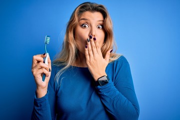 Young beautiful blonde woman using teeth brush over isolated blue background cover mouth with hand...