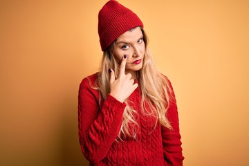 Young beautiful blonde woman wearing casual sweater and wool cap over white background Pointing to the eye watching you gesture, suspicious expression