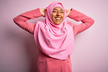 Young African American afro woman wearing muslim hijab over isolated pink background relaxing and stretching, arms and hands behind head and neck smiling happy