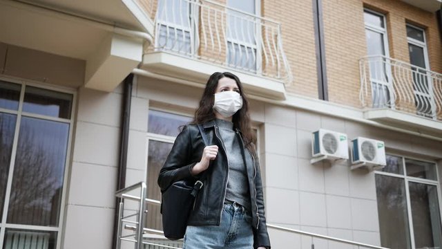 young woman in protective medical mask stands near entrance of house, she is afraid to go around city due to pandemic coronavirus