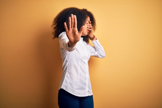 Young beautiful african american elegant woman with afro hair standing over yellow background covering eyes with hands and doing stop gesture with sad and fear expression. Embarrassed and negative