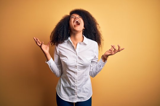 Young beautiful african american elegant woman with afro hair standing over yellow background crazy and mad shouting and yelling with aggressive expression and arms raised. Frustration concept.