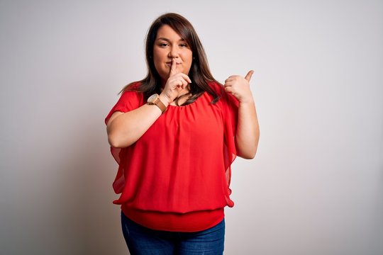 Beautiful brunette plus size woman wearing casual red t-shirt over isolated white background asking to be quiet with finger on lips pointing with hand to the side. Silence and secret concept.