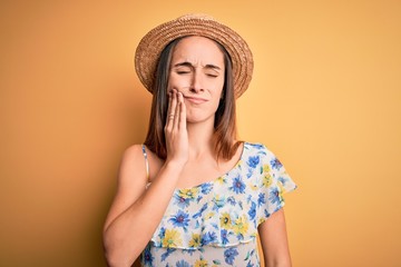 Young beautiful woman wearing casual t-shirt and summer hat over isolated yellow background touching mouth with hand with painful expression because of toothache or dental illness on teeth. Dentist