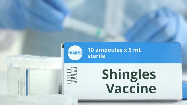 Box with shingles vaccine on the table against blurred lab assistant or doctor. Fictional phaceutical logo