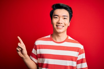 Young handsome chinese man wearing casual striped t-shirt standing over red background with a big smile on face, pointing with hand finger to the side looking at the camera.
