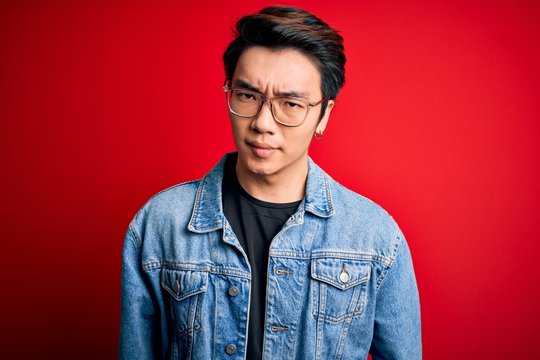 Young handsome chinese man wearing denim jacket and glasses over red background skeptic and nervous, frowning upset because of problem. Negative person.