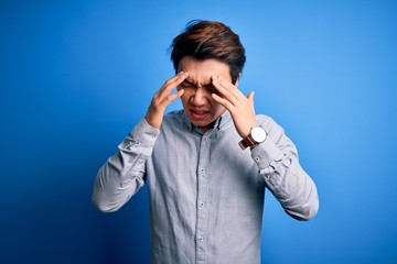 Young handsome chinese man wearing casual shirt standing over isolated blue background with hand on headache because stress. Suffering migraine.