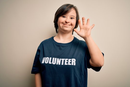 Young down syndrome volunteer woman wearing social care charity t-shirt Waiving saying hello happy and smiling, friendly welcome gesture