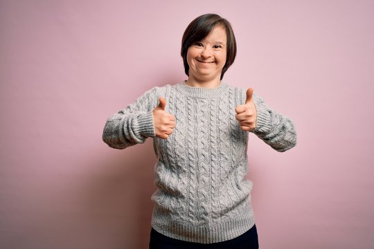 Young down syndrome woman wearing casual sweater over isolated background success sign doing positive gesture with hand, thumbs up smiling and happy. Cheerful expression and winner gesture.