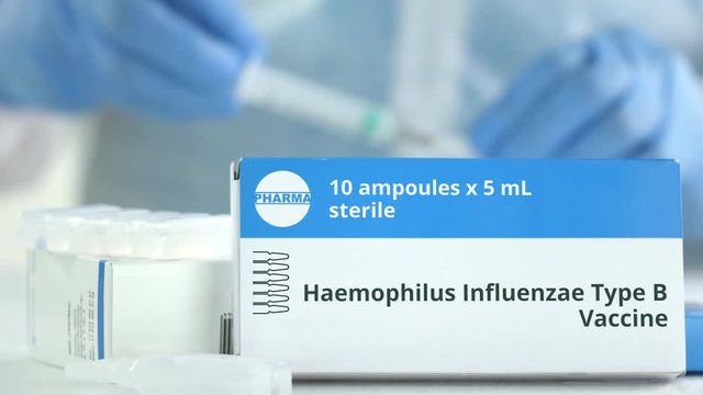 Box with haemophilus influenzae type B vaccine on the table against blurred lab assistant. Fictional phaceutical logo