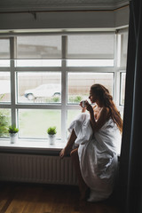 Young woman covered with blanket sitting on windowsill and enjoying hot morning coffee. Cold and rainy autumn weather outside.