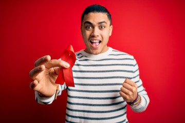 Young brazilian man holding red hiv ribbon standing over isolated background screaming proud and...