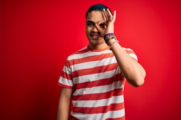 Young brazilian man wearing casual striped t-shirt standing over isolated red background doing ok gesture with hand smiling, eye looking through fingers with happy face.