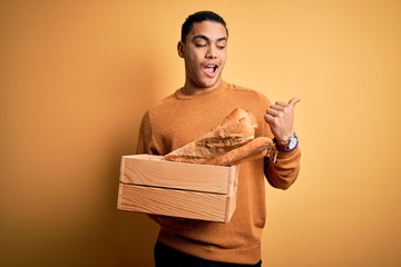 Young baker brazilian man holding box with homemade bread over isolated yellow background pointing and showing with thumb up to the side with happy face smiling