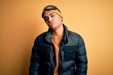 Young brazilian skier man wearing snow sportswear and ski goggles over yellow background looking at the camera blowing a kiss on air being lovely and sexy. Love expression.