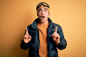 Young brazilian skier man wearing snow sportswear and ski goggles over yellow background amazed and surprised looking up and pointing with fingers and raised arms.