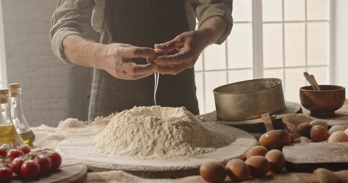 Experienced mature chef cracking an egg into flour to make bread according to traditional recipe. Old man baking at home, enjoying hobby - closeup shot 4k footage