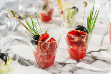 Delicious appetizers in glass cups with prosciutto, olives and tomatoes on marble banquet table. Catering food, canape and snacks, close up