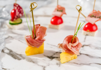 Delicious appetizers with prosciutto and pineapple on marble banquet table. Catering food, canape and snacks, close up