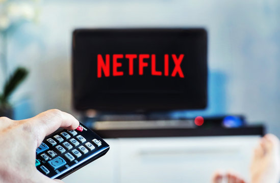 A young man watches Netflix on his TV and at home. TV remote in the foreground, blurred background on the TV.
