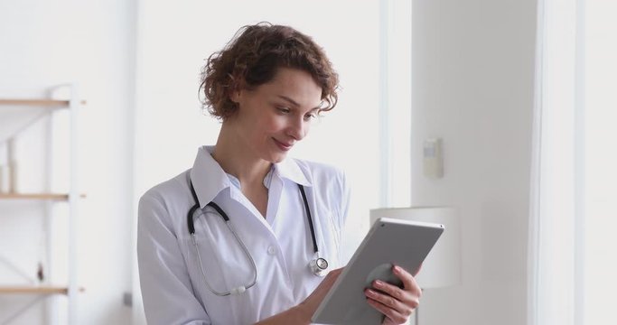 Smiling female physician using digital tablet computer app standing in hospital office. Young woman professional doctor holding touchscreen pad device in clinic. Online healthcare medical tech concept