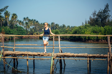 Fototapeta na wymiar A female tourist in denim shorts with short haircut on sunny tropical day. Young pretty girl stands on wooden bridge over small river against palm jungle and blue sky. Trips to picturesque places.