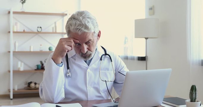 Puzzled senior doctor therapist looking at laptop analyzing data thinking of wrong diagnosis. Concerned serious professional older adult physician doubting on test results working in medical office.
