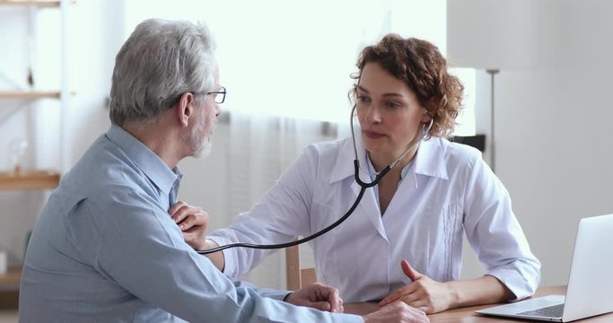 Female doctor holding stethoscope examining old senior 60s grandpa patient in hospital. Cardiologist checking elderly man heartbeat at checkup medical visit. Older people cardiology healthcare concept