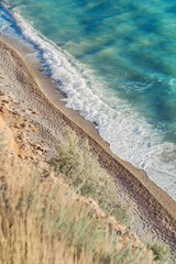 Sunny coastline, beach. View from above on sand, blue sea water and white foamy waves. 