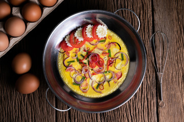 Steamed eggs with chili, spring onion, cucumbers and tomato in a pot and eggs in a panel on a wooden floor.
