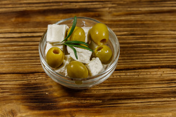 Feta cheese cubes with green olives and rosemary in glass bowl on a wooden table