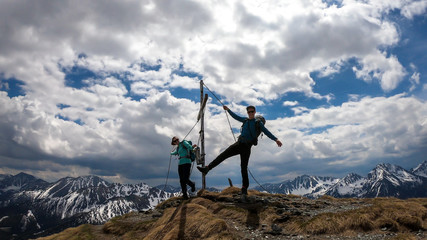 A couple with hiking backpacks standing on the top of Himmeleck in Austrian Alps. There is a massive mountain range in the back, partially covered with snow. Having fun, playtime. Cross on the top