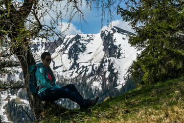 A woman in hiking outfit sitting under a huge tree and enjoying the Alpine landscape, while hiking to the Himmeleck peak. High mountains covered with snow. Early spring in Alps. Freedom and adventure