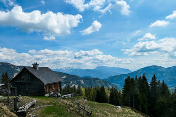 Fototapeta na wymiar A small cottage located at the mountain ledge with the vast, panoramic view on the Alps in Austria. There are many mountain ranges in the back. Spring coming to the Alps. Dense forest
