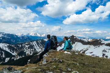 Couple with big hiking backpacks resting on their way to Himmeleck, Austrian Alps and enjoying the view. There is a massive mountain range in the back, partially covered with snow. Early spring vibes