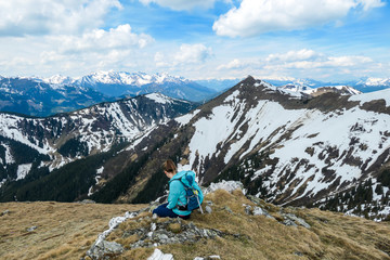 Fototapeta premium A woman with big hiking backpack resting on her way to Himmeleck, Austrian Alps and enjoying the view. There is a massive mountain range in the back, partially covered with snow. Early spring vibes