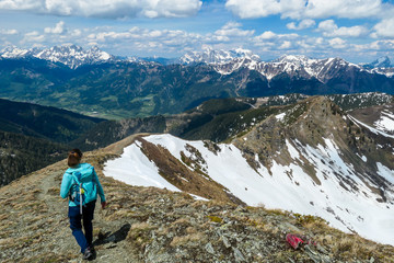 Fototapeta premium A woman with big hiking backpack hiking to Himmeleck peak, Austrian Alps. There is a massive mountain range in the back, partially covered with snow. Early spring vibes. Freedom and adventure.