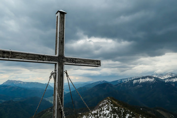 Fototapeta na wymiar Close up on wooden cross at the top of Himmeleck, Austrian Alps. There is a massive mountain range in the back, partially covered with snow. Early spring. barren mountain slopes. Overcast. Achievement
