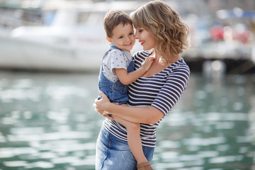 Fototapeta na wymiar Mother and little son spend time outdoors in the summer together, sitting on a pier near boats and boats.Young mother plays with her baby on the promenade.Summer vacation for two by the sea. Mother an
