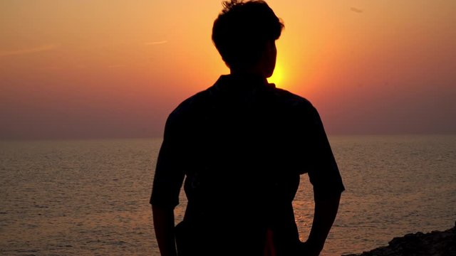 Rear view silhouette of young Asian man traveler walking on island mountain sea cliff and feeling freedom with outstretched his arms in the air at summer sunset with twilight sky in summer vacation.