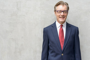 Happy handsome mature businessman in suit smiling against concrete wall