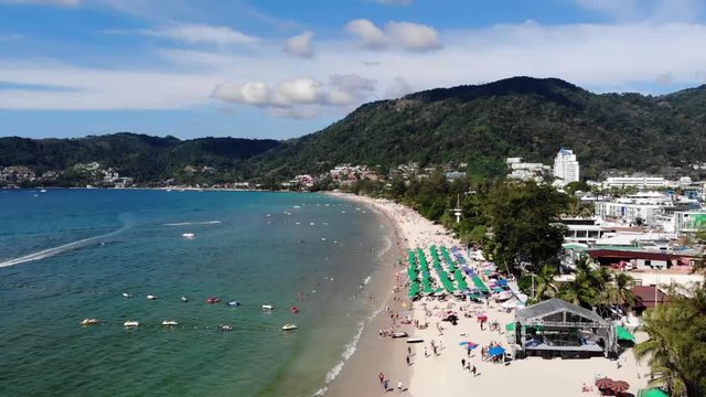 Aerial views overlooking the white sand Patong Beach in Phuket, with a jet ski, parasailing, and mountains in the background. Pedestal up shot.