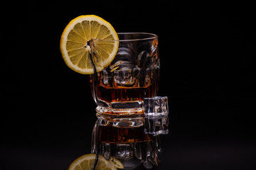 A glass of whiskey with pieces of ice and lemon stands on a black glass in which its reflection is visible.