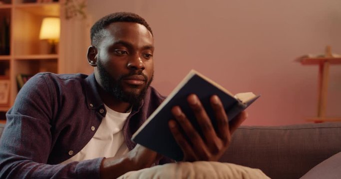 Close up of carefree handsome guy turning page while reading book. Young bearded man enjoying free time while sitting on sofa at home. Concept of leisure and hobby.