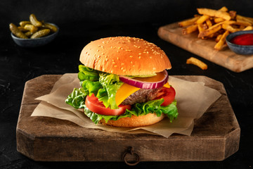 Burger with beef, cheese, onion, tomato, and green salad, on a dark background with pickles and French fries