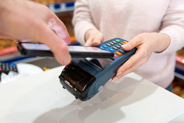 A woman's hands are held by a payment terminal and a man pays for a purchase using a smartphone. Close up. The concept of NFC, business and banking transactions
