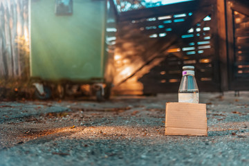 The concept of environmental pollution and waste sorting. A glass bottle stands on the ground in front of a dumpster. Near cardboard with empty blank. Copy space
